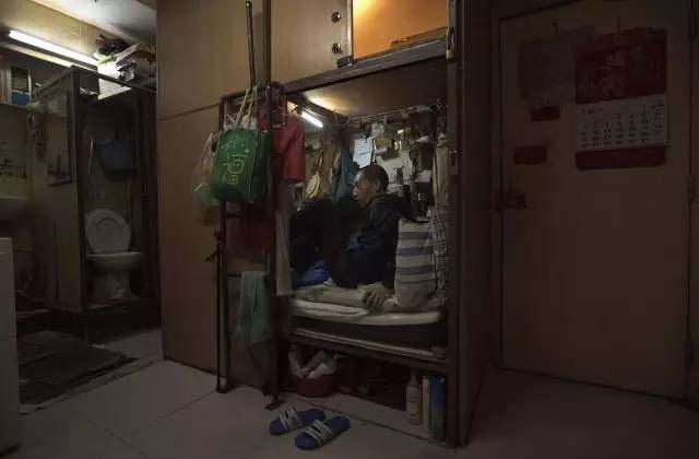 黄达明，63岁，他正蜷缩着双腿坐在自己的“棺材屋”内。为了这间房子，他每个月要支付2400港币(约人民币2100元)。左侧的简易厕所由这层的所有租客共用