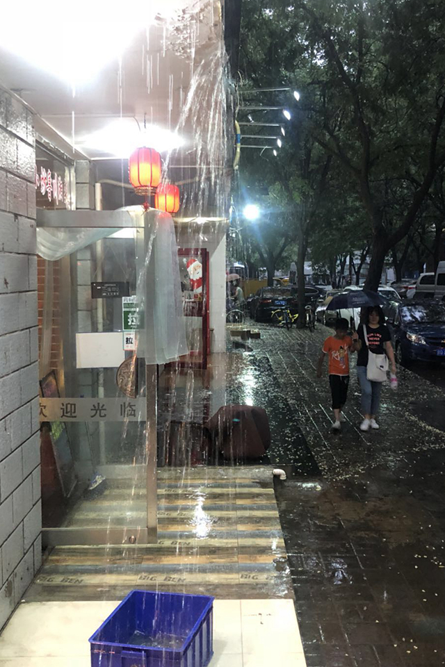 小店门口雨水向瀑布一样顺墙头留下，一对母子撑伞路过。