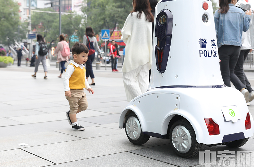 9月27日，在北京西单商业区，一台机器人“警察”出现在来往的人群中“上岗执勤”，吸引了众多市民驻足。记者在现场看到，这台机器人上装有摄像头，扬声器，照明灯，报警灯和轮子等，显得科幻风十足。据了解，这款警用机器人是由公安部第一研究所研制。图为机器人吸引了一名小朋友的注意。 中宏网记者 富宇 摄
