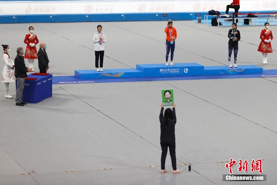 图为女子3000米比赛颁奖仪式中，引导员用提示牌指导运动员佩戴和摘下口罩拍照。 中新社记者 富田 摄